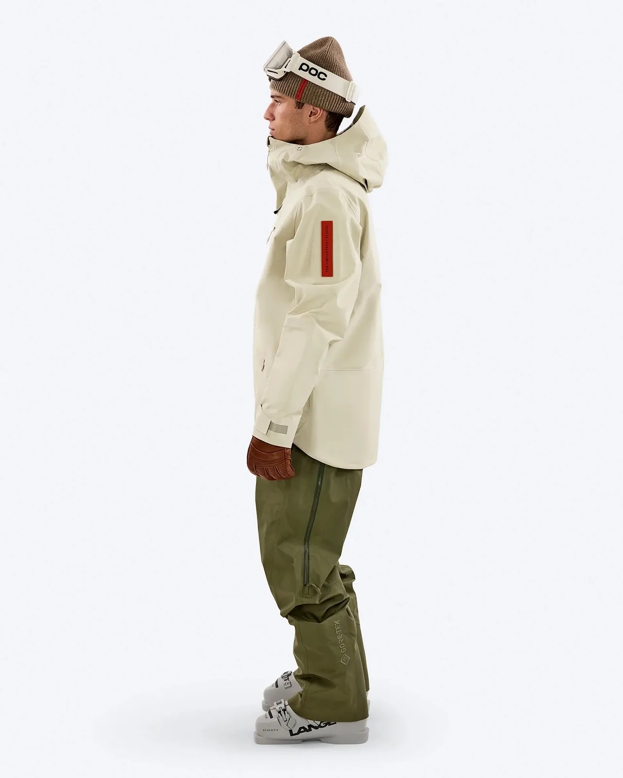 Mann in Skikleidung: Beiger Anorak mit Kapuze, grüne Hose, Skischuhe und braune Handschuhe, trägt Mütze und Skibrille.