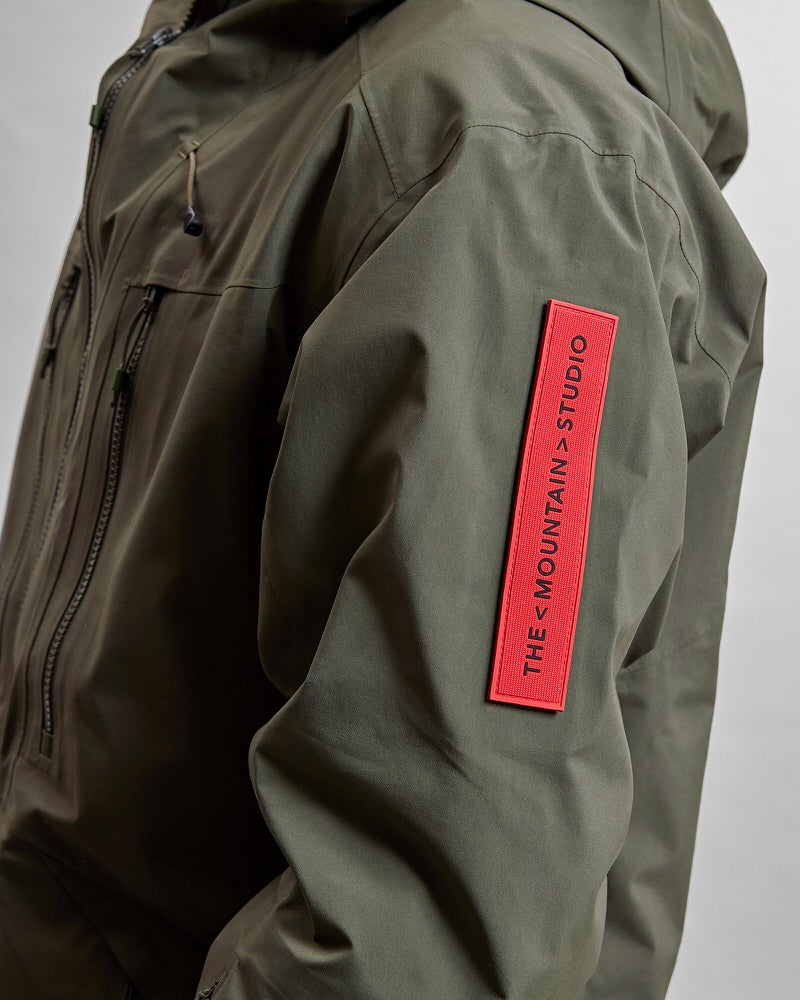 Detailaufnahme einer grünen Z-2 HD GORE-TEX Pro 3L Shell Jacket mit rotem "THE MOUNTAIN STUDIO" Aufnäher am Ärmel. Reissverschlüsse sichtbar.
