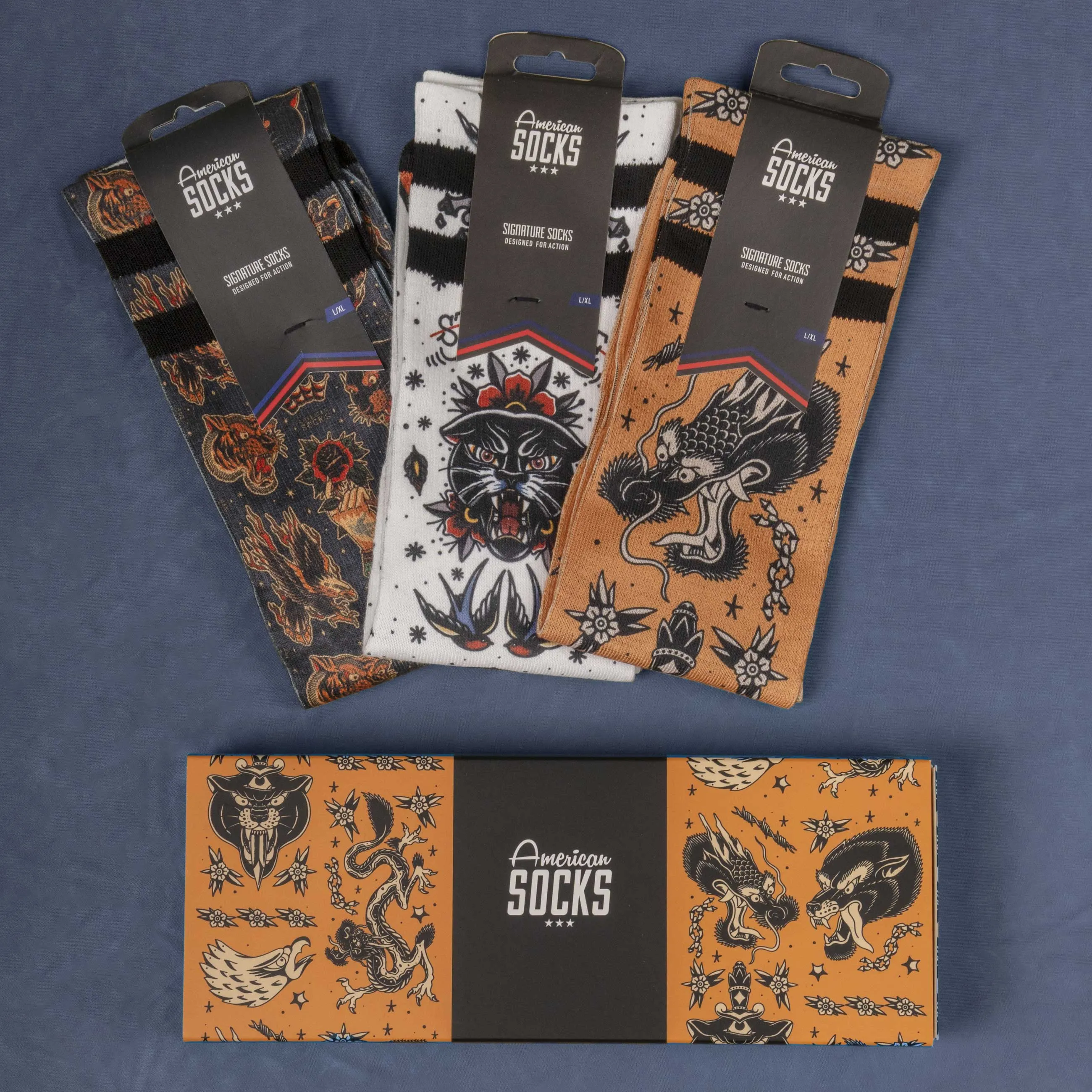Drei verpackte American Socks mit Tattoo-Motiven und eine passende Geschenkbox liegen auf einem blauen Untergrund.