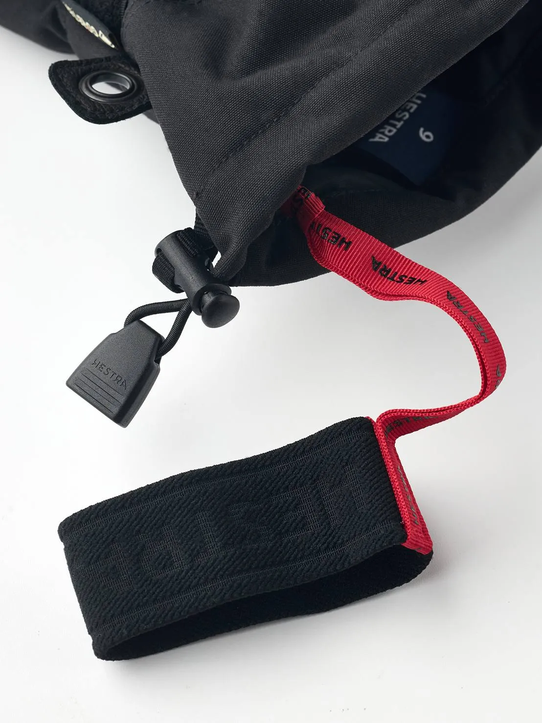 Detailaufnahme eines schwarzen Hestra Gore-Tex Fünffinger-Handschuhs mit roter Schlaufe und schwarzem Verschluss.