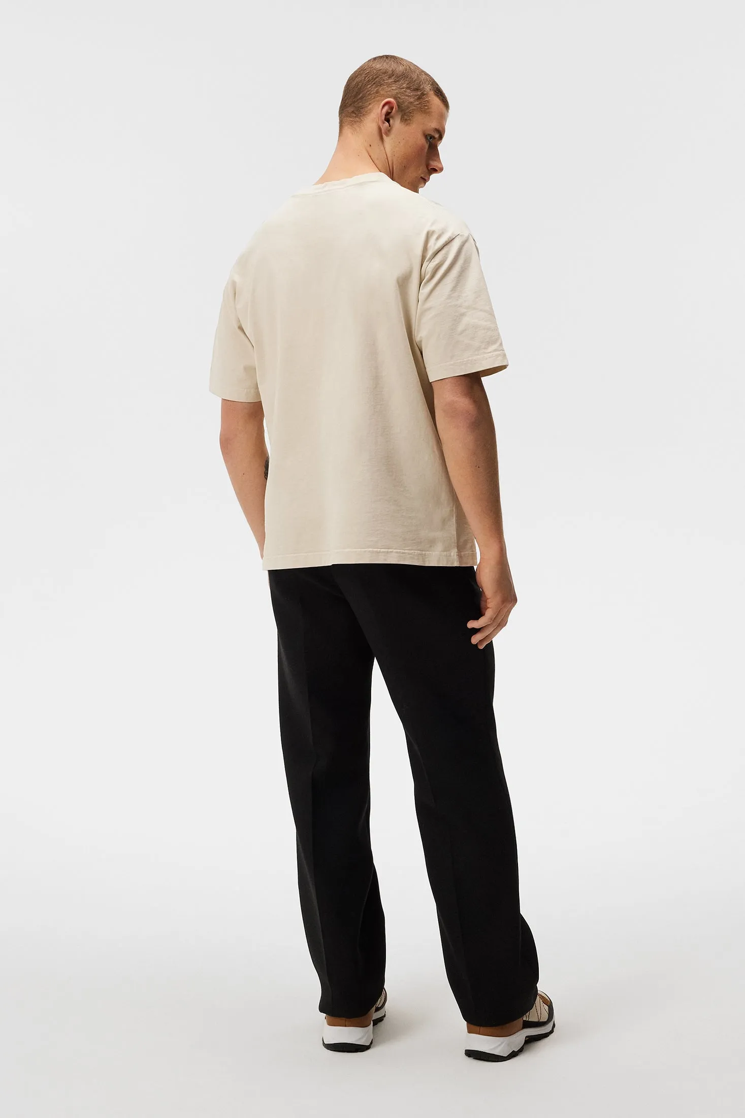 Ein Mann trägt ein Oyster Gray Walsh Boxy Print Tee, kombiniert mit schwarzen Hosen, von hinten auf einem weißen Hintergrund.