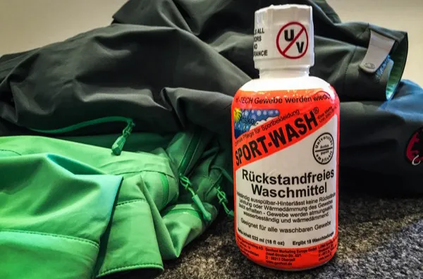 Grüne und graue Jacke, neben einer Flasche Sportwaschmittel auf grauem Untergrund.