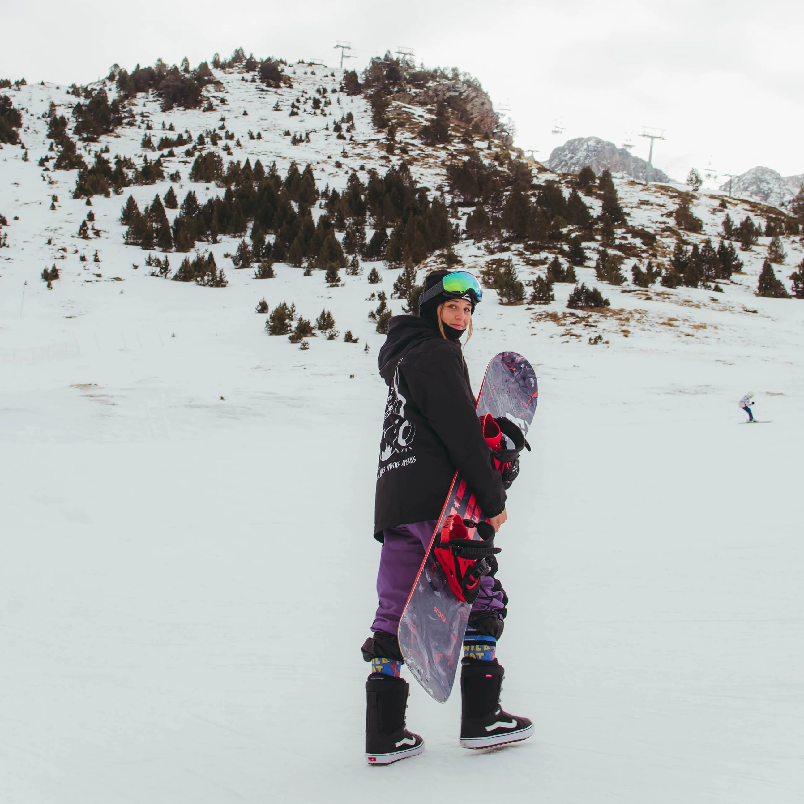 Snowboarderin mit Snowboard vor verschneiter Berglandschaft, trägt Wild Nature Snow Socks.