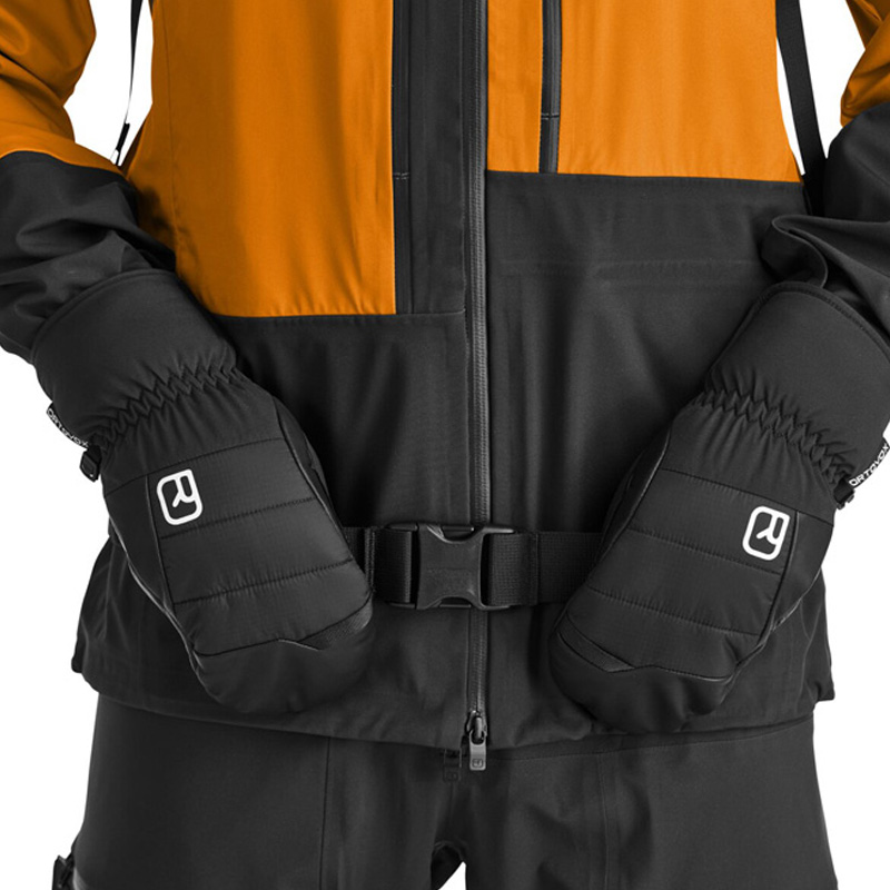 Schwarze Fäustlinge Detail Detailaufnahme: Schwarze Ortovox Freeride Cozy Fäustlinge vor einer Winterjacke mit Gürtel.