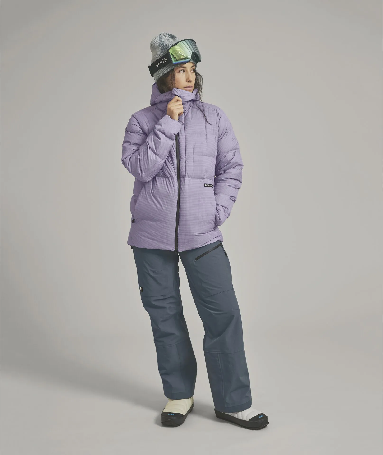 Eine Frau in lila Daunenjacke, grauer Mütze und Skibrille blickt auf einem hellen Hintergrund nach rechts. Sie trägt blaue Hosen und Hausschuhe.