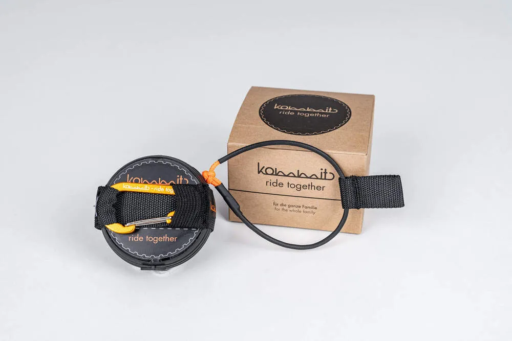 Das kommit Zugsystem: Eine schwarze Spule mit Gurtband und Karabiner liegt neben einer braunen Schachtel. Schwarzes Seil verbunden.