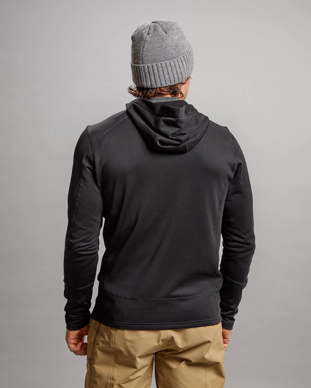 Rückansicht einer schwarzen M-4 Techno Stretch Mid Hood Jacke mit Kapuze, kombiniert mit einer grauen Mütze und beigen Hosen.