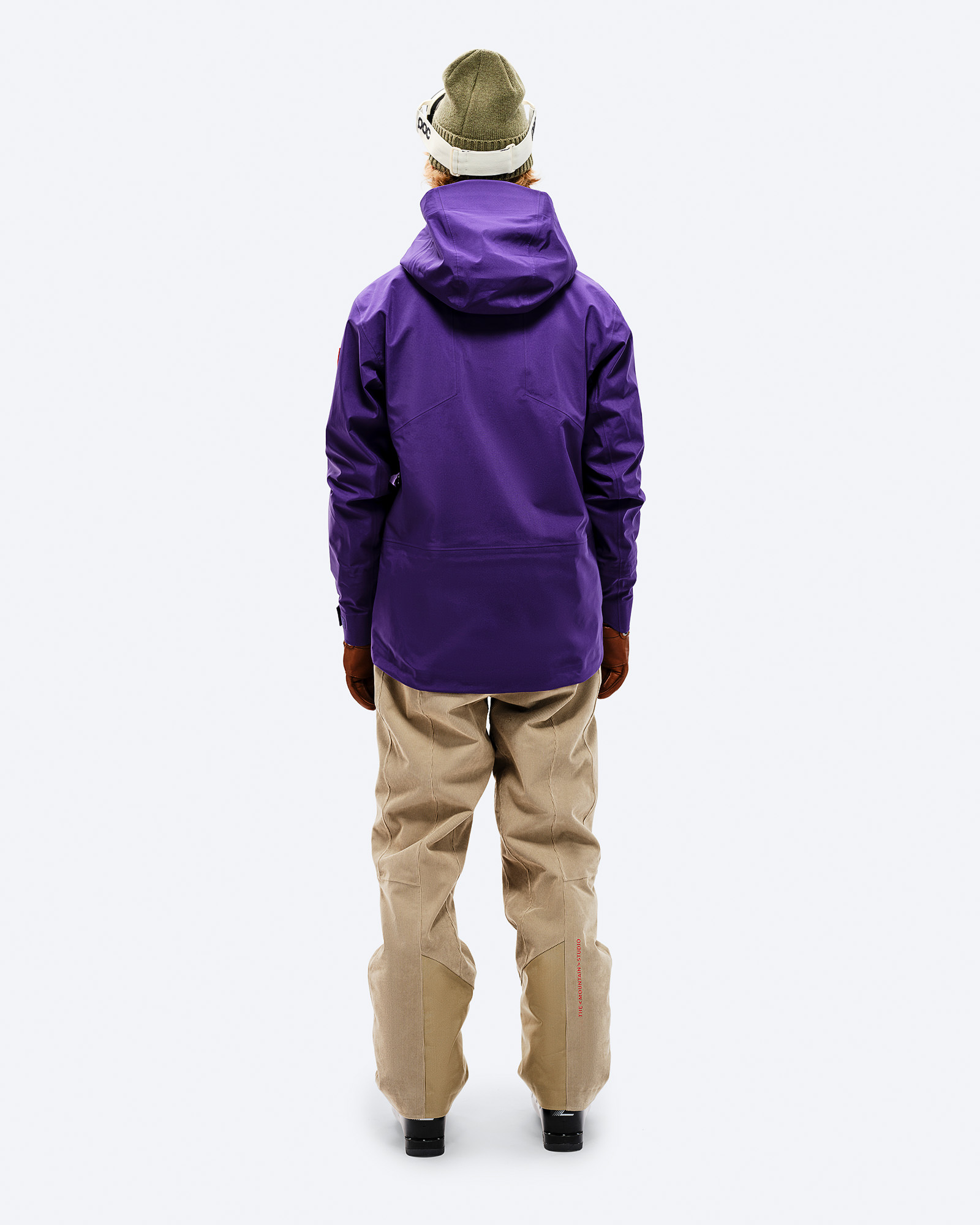 Person von hinten in lila GORE-TEX Shell Jacket, beigen Hosen, Mütze und Skibrille.