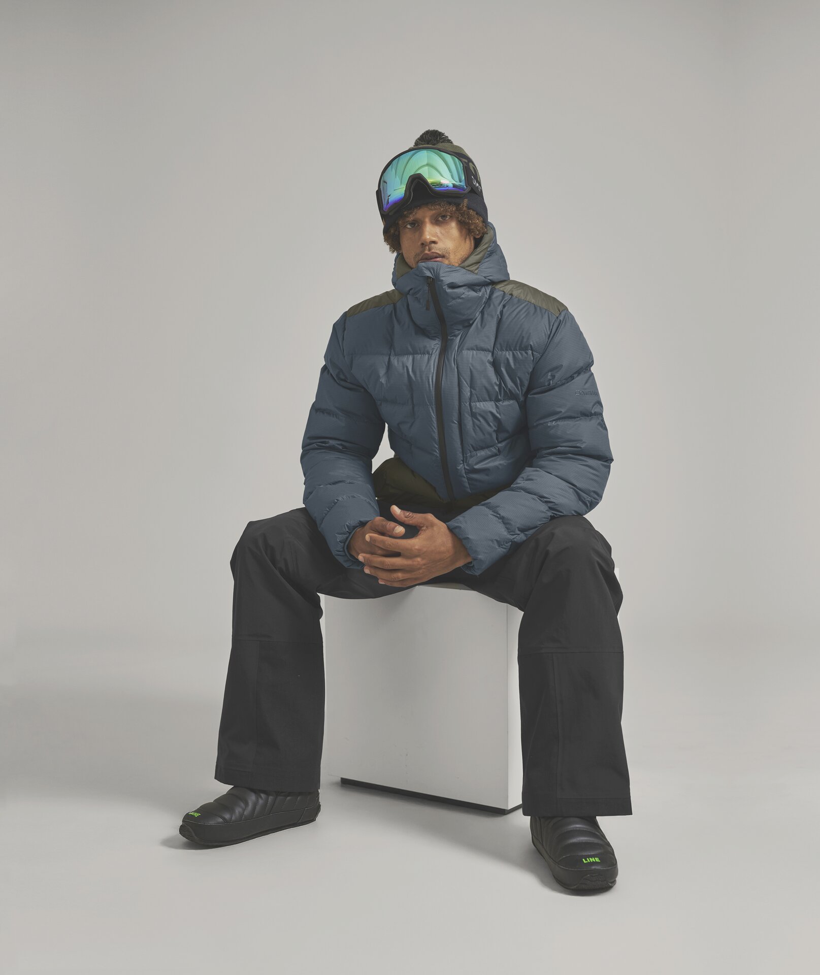 Winterlicher Mann auf Würfel Mann mit Skibrille, Mütze und Winterkleidung sitzt auf einem weißen Würfel. Er trägt eine blaue Daunenjacke, schwarze Hose und schwarze Schuhe.