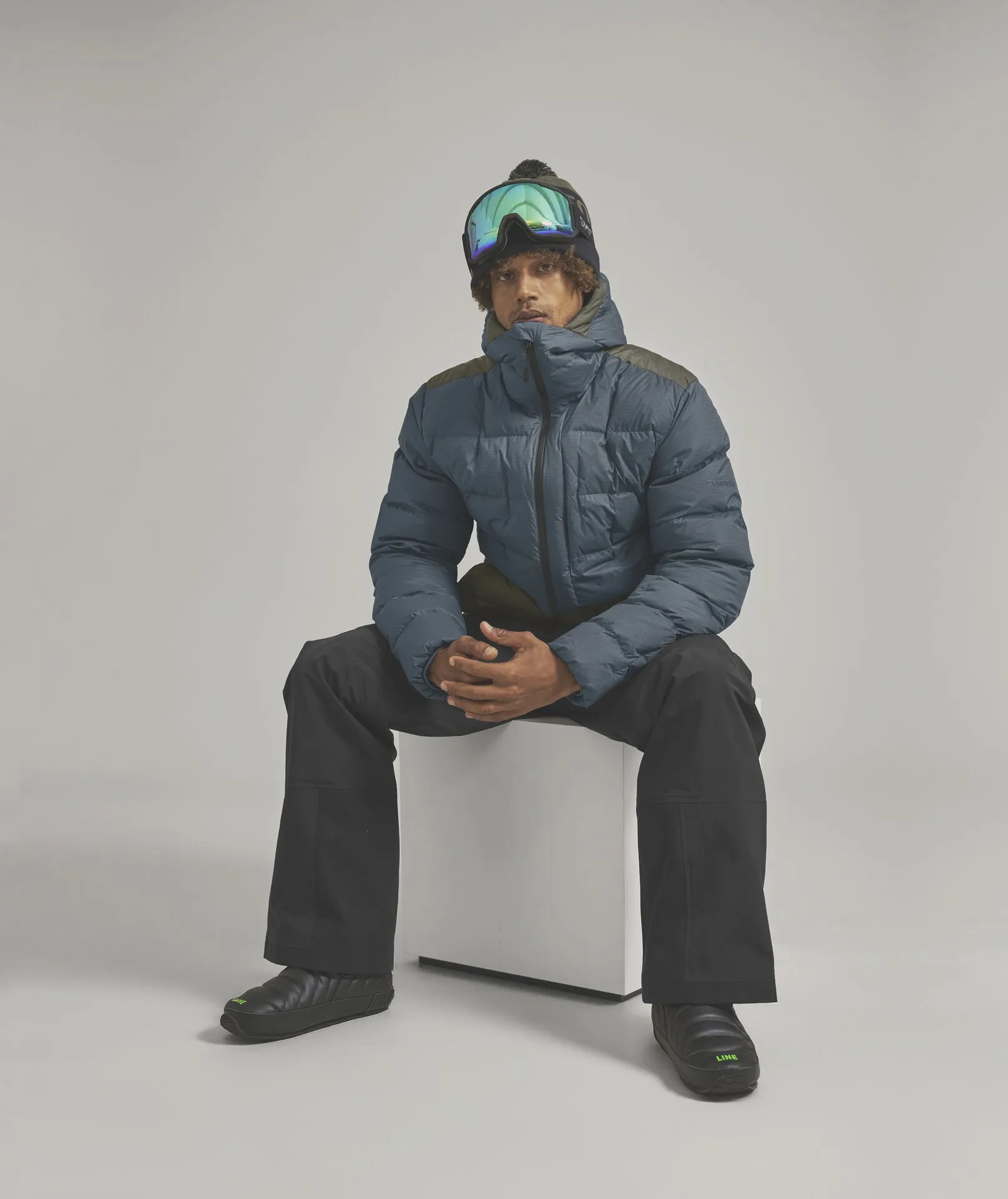 Mann mit Skibrille, Mütze und Winterkleidung sitzt auf einem weißen Würfel. Er trägt eine blaue Daunenjacke, schwarze Hose und schwarze Schuhe.