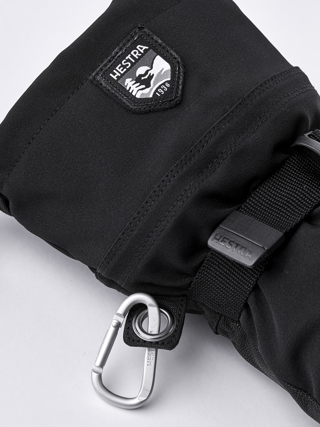 Nahaufnahme eines schwarzen Hestra Army Leather Extreme Mitt Fäustlings mit Karabiner und Logo.