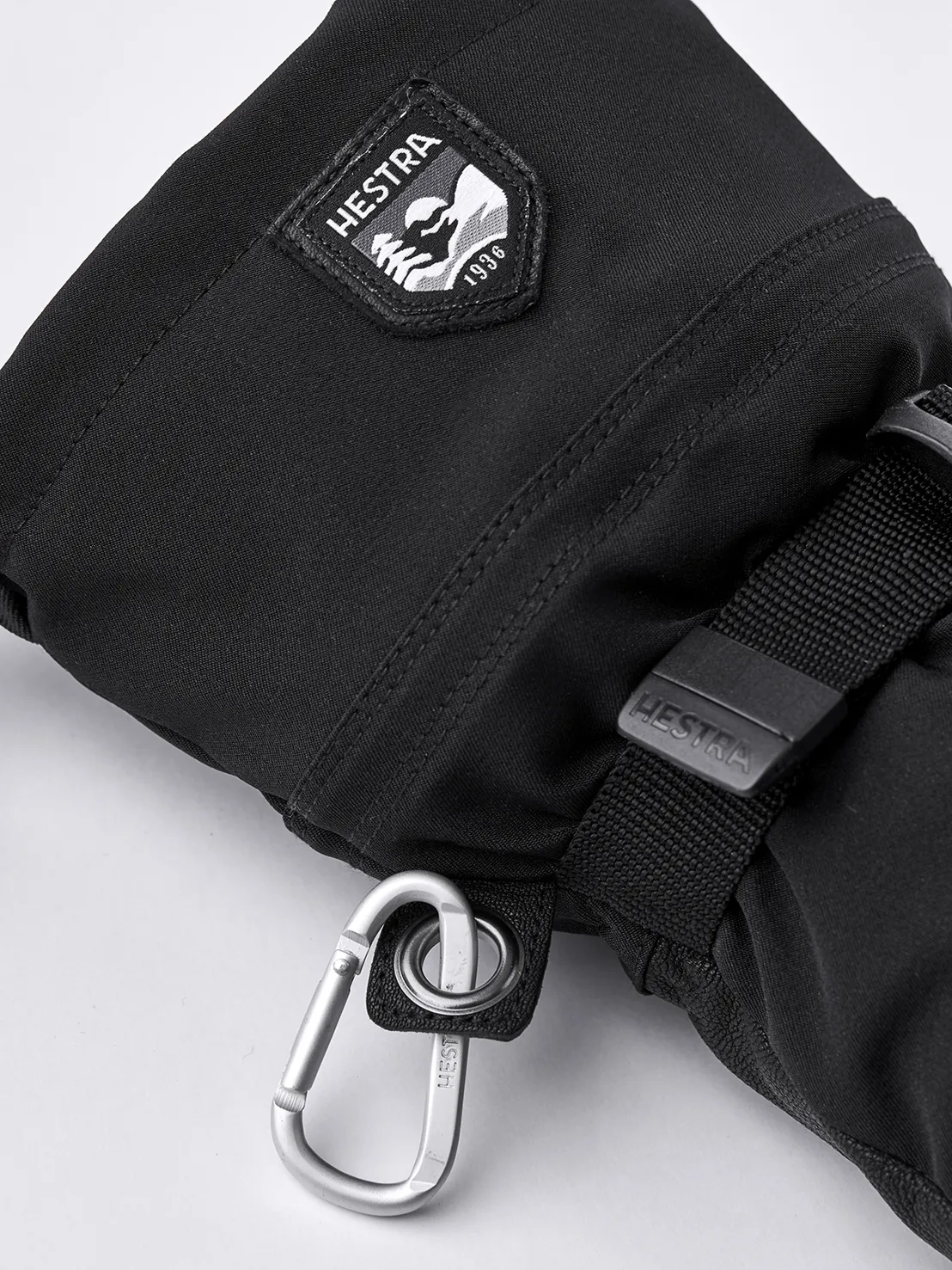 Nahaufnahme eines schwarzen Hestra Army Leather Extreme Mitt Fäustlings mit Karabiner und Logo.