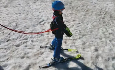 Kind mit Ski Trainer auf Schnee. Das Kind trägt einen blauen Helm und wird mit Leinen geführt.