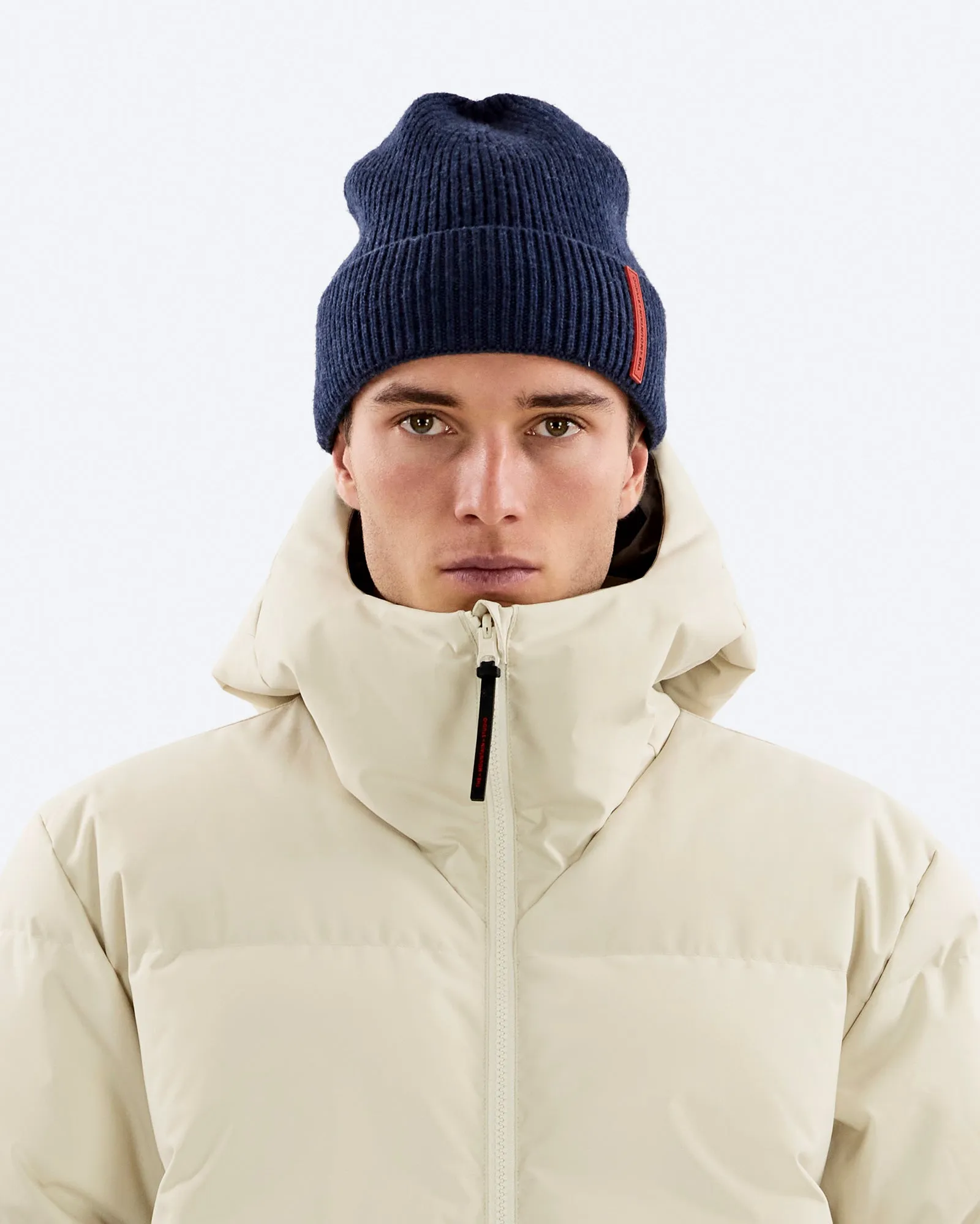 Porträt eines Mannes mit blauer Mütze und beiger Daunenjacke vor einem weißen Hintergrund.