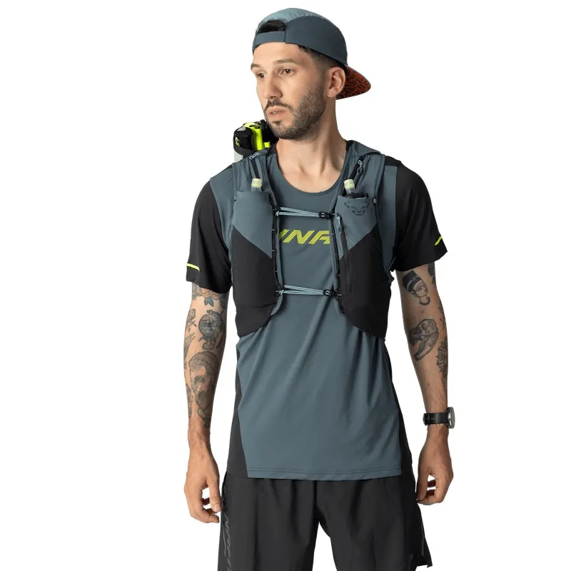 Mann in Sportbekleidung mit Tätowierungen trägt Laufweste. Graue Kappe, Blick nach links. Grau-schwarze Weste mit Taschen für Trinkflaschen. Graues T-Shirt mit gelbem "DYNAFIT"-Aufdruck. Uhr links. Weißer Hintergrund.