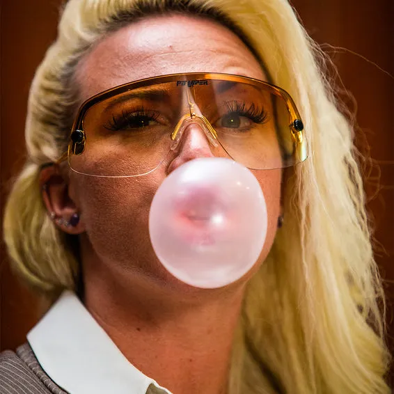 Blonde Frau mit Sonnenbrille und Kaugummi-Blase vor dem Gesicht. Gold Standard Look.