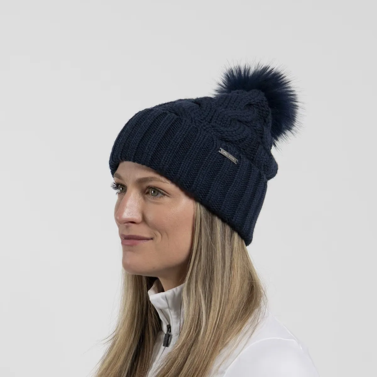 Eine Frau mit langen blonden Haaren trägt eine dunkelblaue Strickmütze mit Bommel und Zopfmuster. Sie blickt nach links.