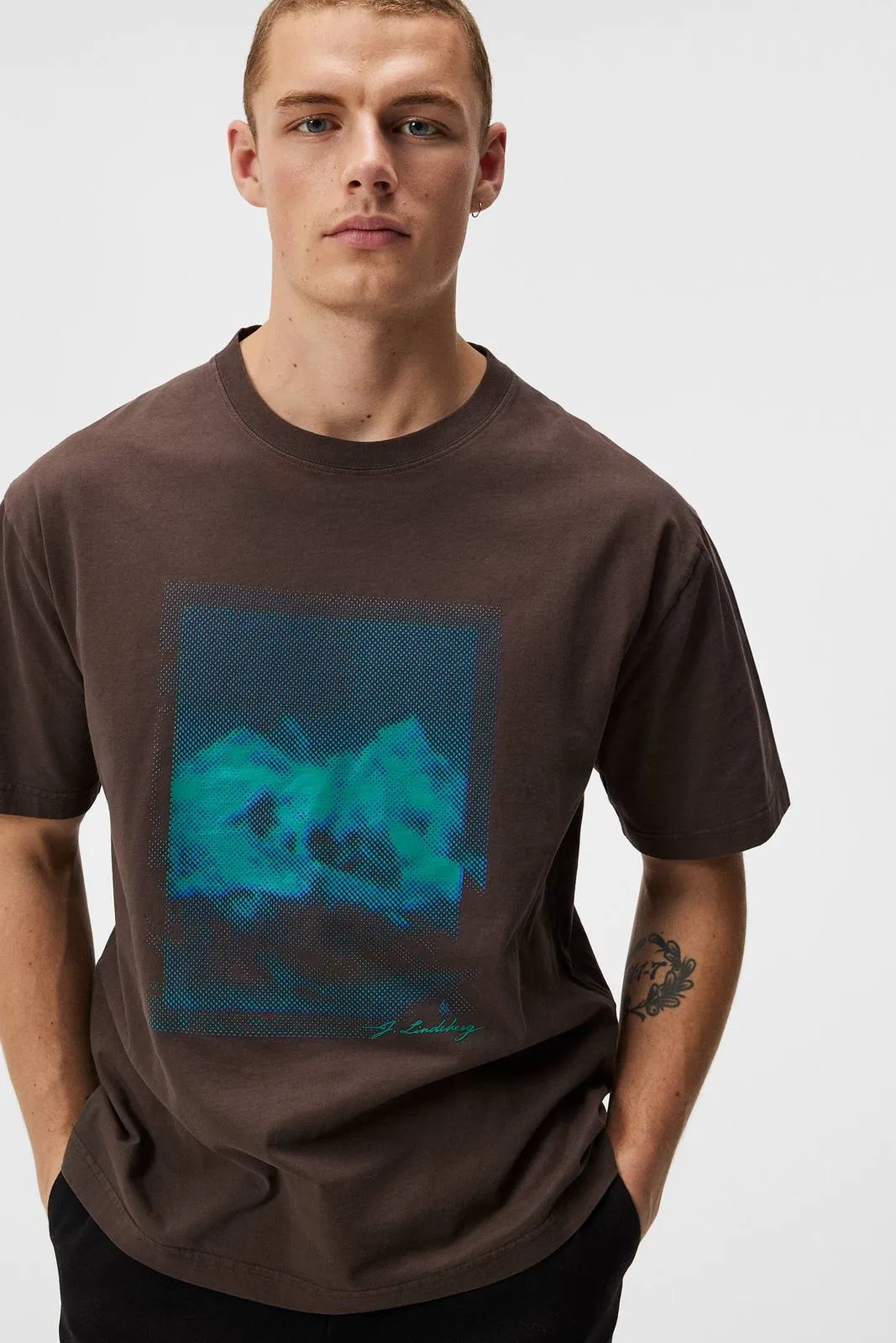 Mann trägt braunes T-Shirt mit blau-grünem Print. Der Print ist ein abstraktes Muster mit dem Schriftzug "J. Lindeberg".