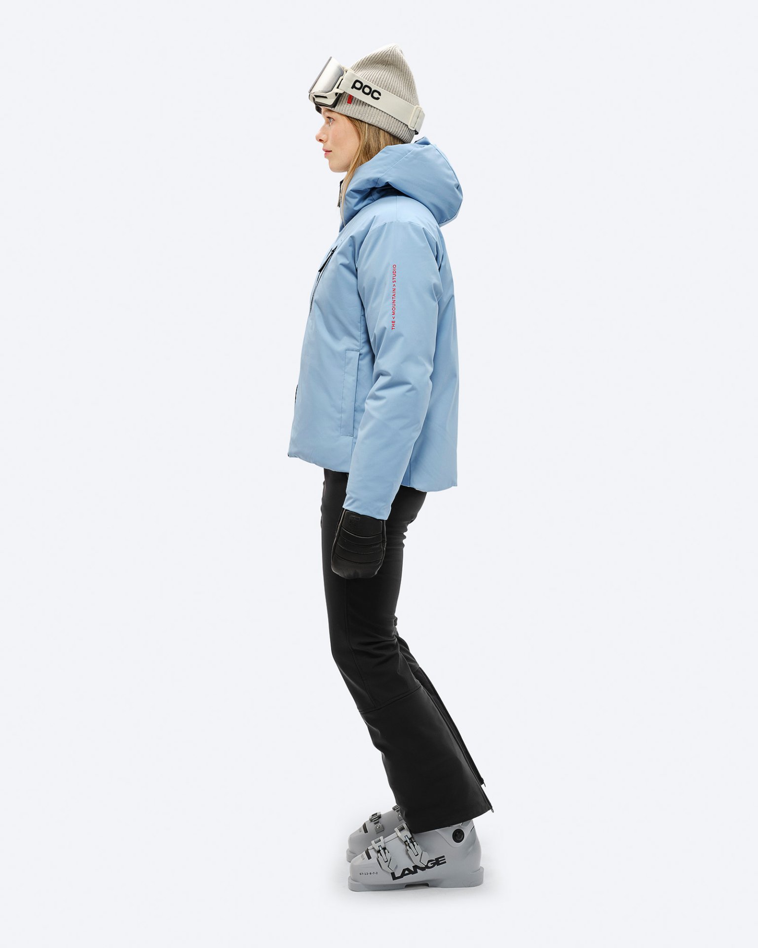 Frau in Skiausrüstung: Hellblaue Daunenjacke, schwarze Hose, Skischuhe, Mütze mit Skibrille, Profilansicht.