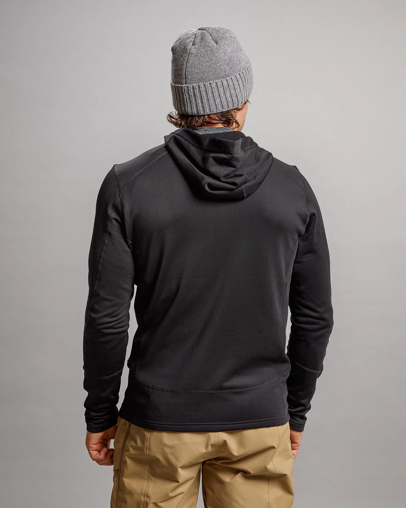 Rückansicht einer schwarzen M-4 Techno Stretch Mid Hood Jacke mit Kapuze, kombiniert mit einer grauen Mütze und beigen Hosen.