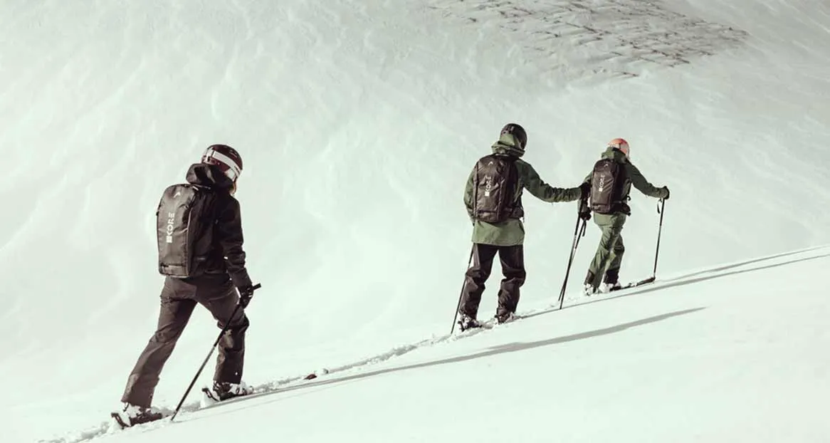 Drei Personen beim Skitourengehen, von hinten gesehen, mit Skiern und Stöcken auf einer verschneiten Piste. Sie tragen Helme und Rucksäcke.