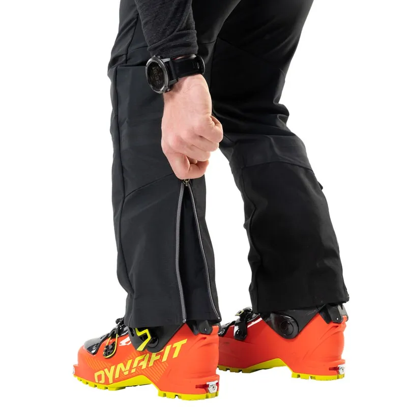 Nahaufnahme: Person öffnet Reißverschluss an schwarzer Radical Infinium Hybrid Hose über Skischuh.