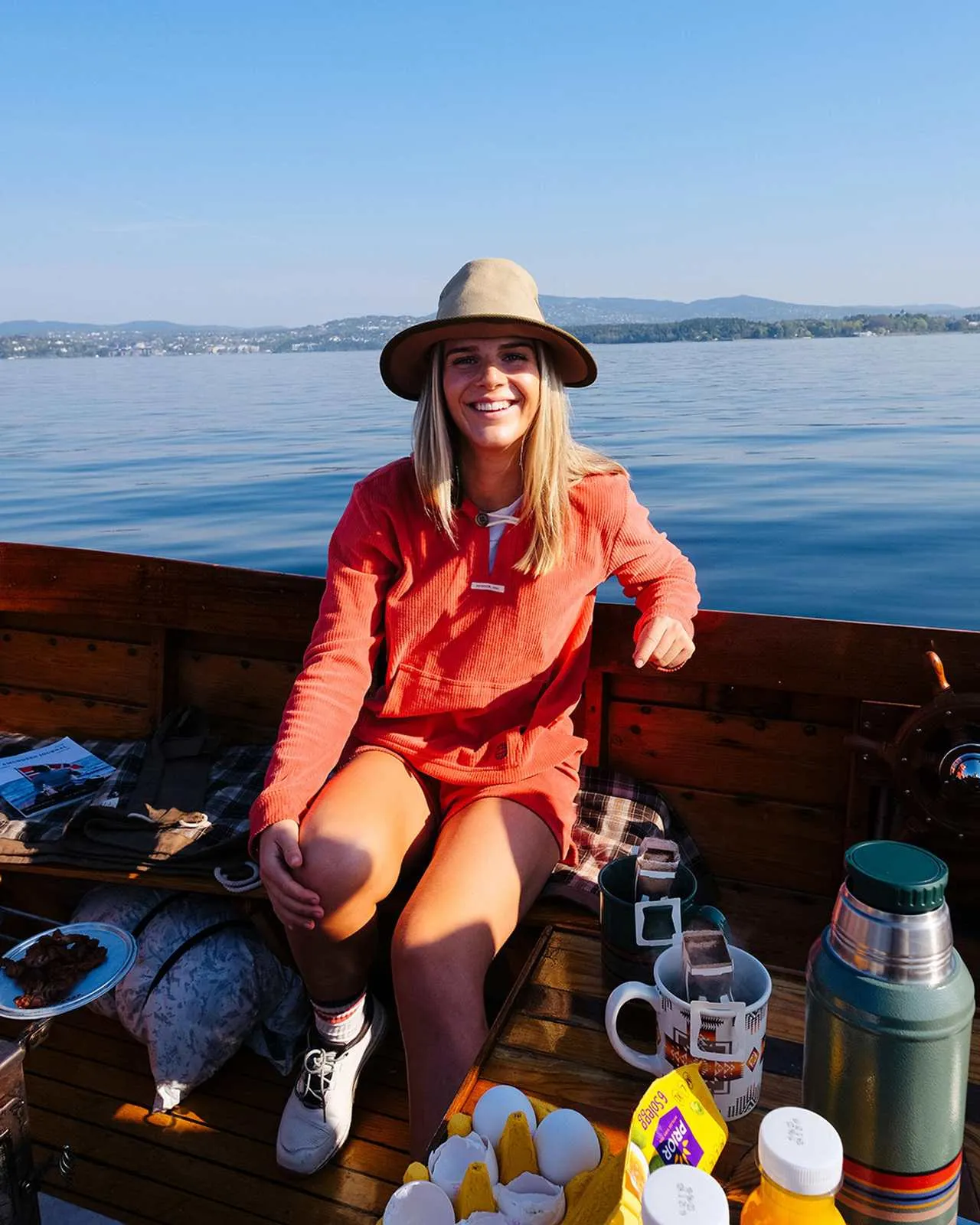 Eine lächelnde Frau mit Hut sitzt auf einem Holzboot vor blauem Wasser und Bergen, umgeben von Speisen und Getränken.