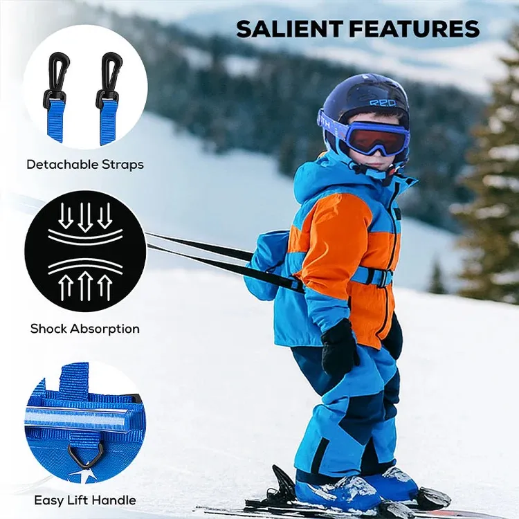 Ein Kleinkind in orange-blauer Skibekleidung, Helm und Skibrille steht auf Skiern auf einem verschneiten Hang. Es trägt ein blaues Skigeschirr mit schwarzen Leinen am Rücken. Details: abnehmbare Gurte, Dämpfung, Hebegriff.