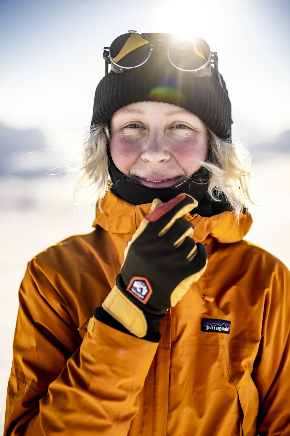 Lächelnde Person mit Mütze und Skibrille auf dem Kopf, trägt orangefarbene Jacke und Handschuhe im Schnee.