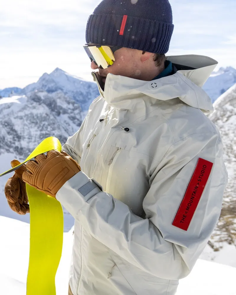 Ein Mann in Z-2 HD GORE-TEX Pro 3L Shell Jacket "Castle Wall" mit Skibrille und Mütze hält Skifell vor verschneiten Bergen.