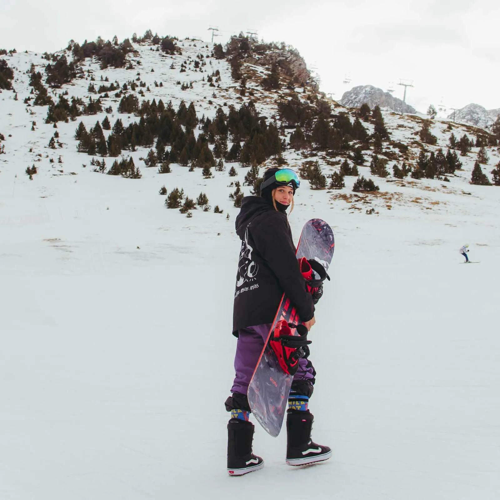 Snowboarderin mit Snowboard vor verschneiter Berglandschaft, trägt Wild Nature Snow Socks.