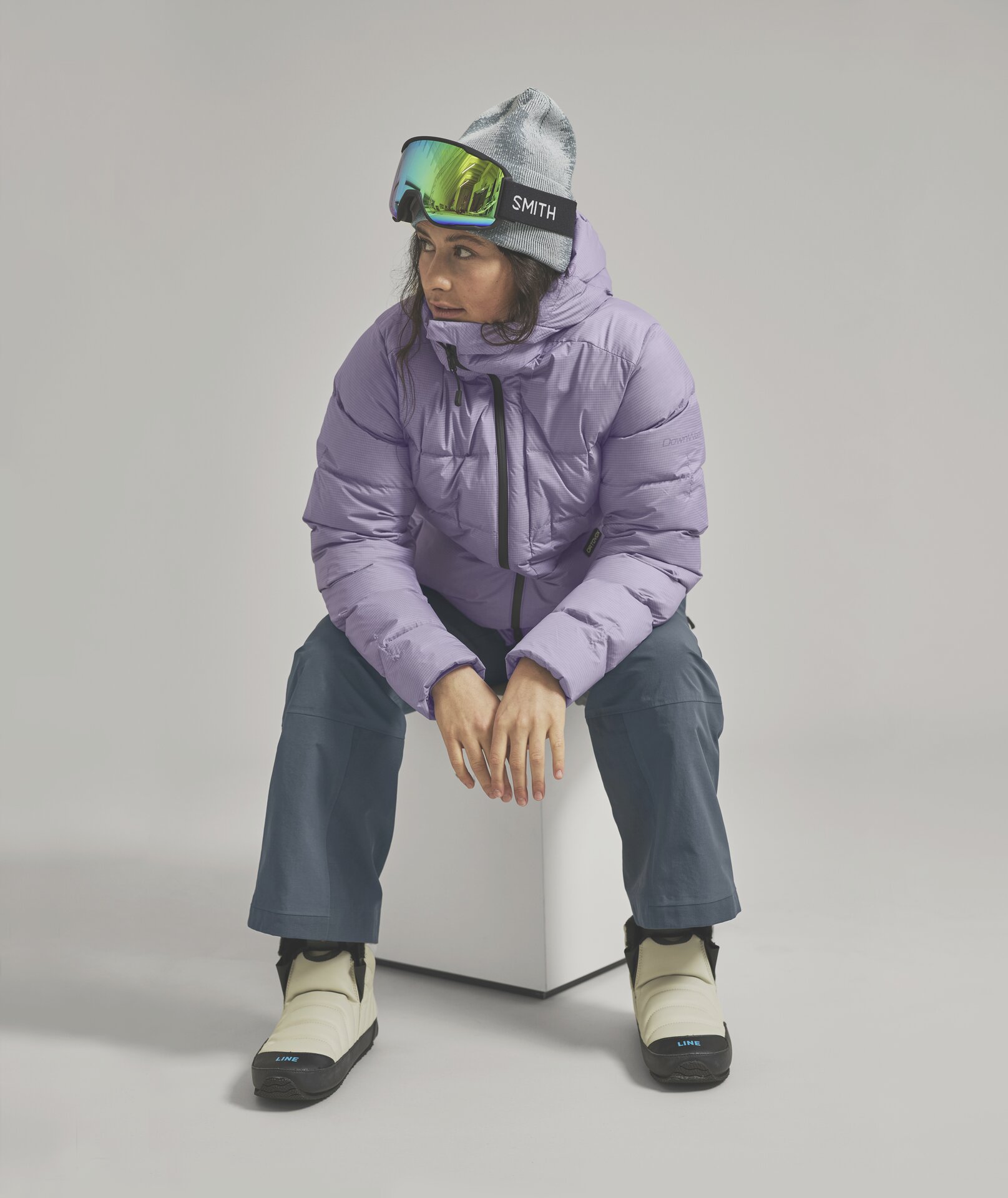 Skifahrerin in lila Jacke Eine Frau mit Mütze und Skibrille, trägt eine lila Daunenjacke, graue Hose und cremefarbene Stiefel, während sie auf einem weißen Würfel sitzt.