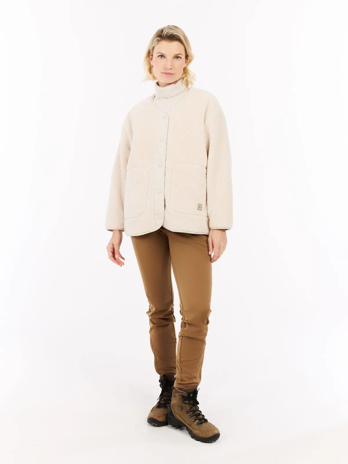 Eine Frau mit blonden Haaren, die eine helle Jacke, braune Hose und Wanderschuhe vor einem weißen Hintergrund trägt.