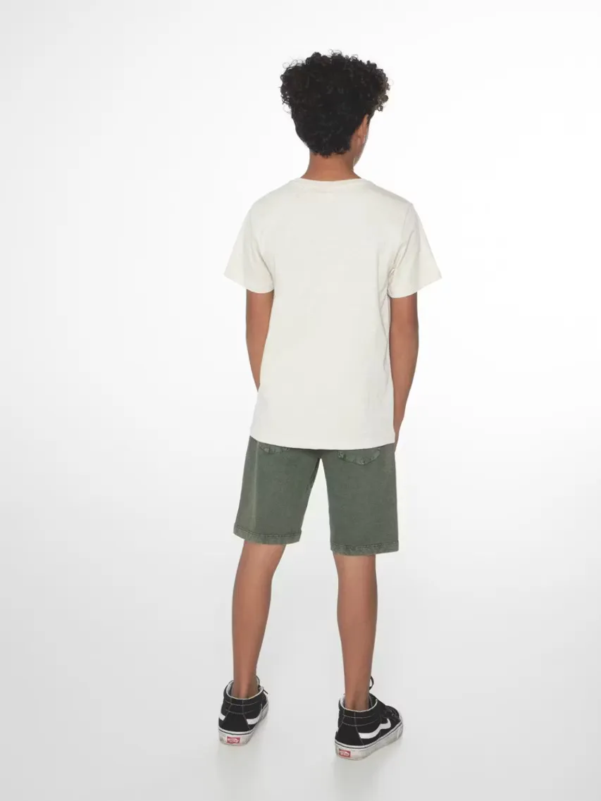 Ein Kind mit lockigem Haar von hinten, bekleidet mit einem cremefarbenen T-Shirt und olivgrünen Shorts. Die Schuhe sind schwarz-weiß gemusterte Sneaker. Das Kind steht vor einem hellen Hintergrund.