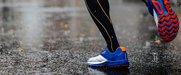 Ein Läufer in Sportschuhen und Socken läuft auf nassem Asphalt. Die Sohle des Schuhs ist rot und blau.
