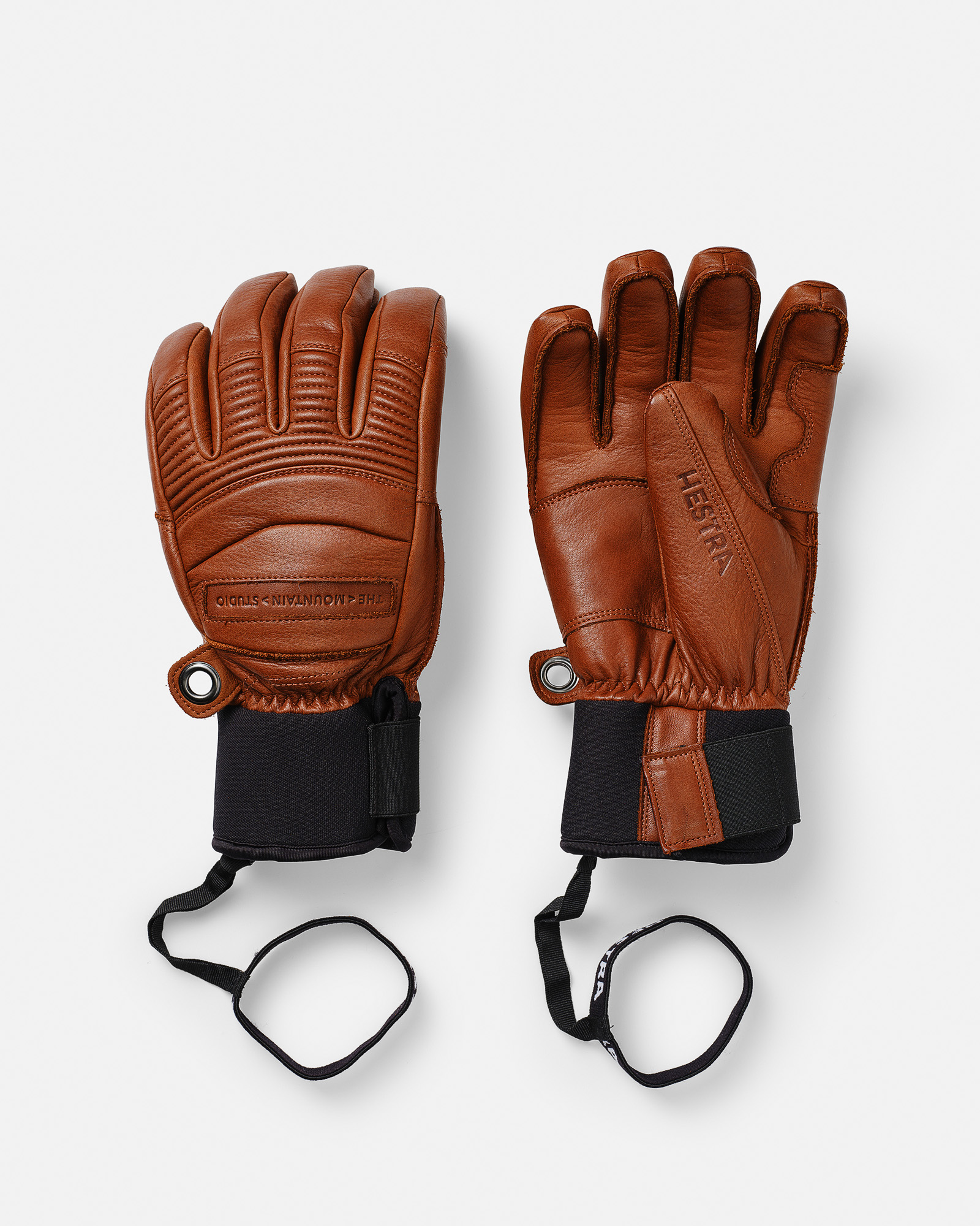 Zwei braune HESTRA Lederhandschuhe mit schwarzen Handgelenkbändern auf weißem Grund.